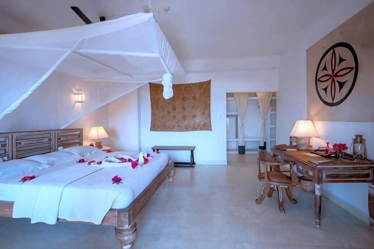 Quarto de hotel com cama, mesa de cabeceira, luminária e área de refeições com cadeiras e mesa.