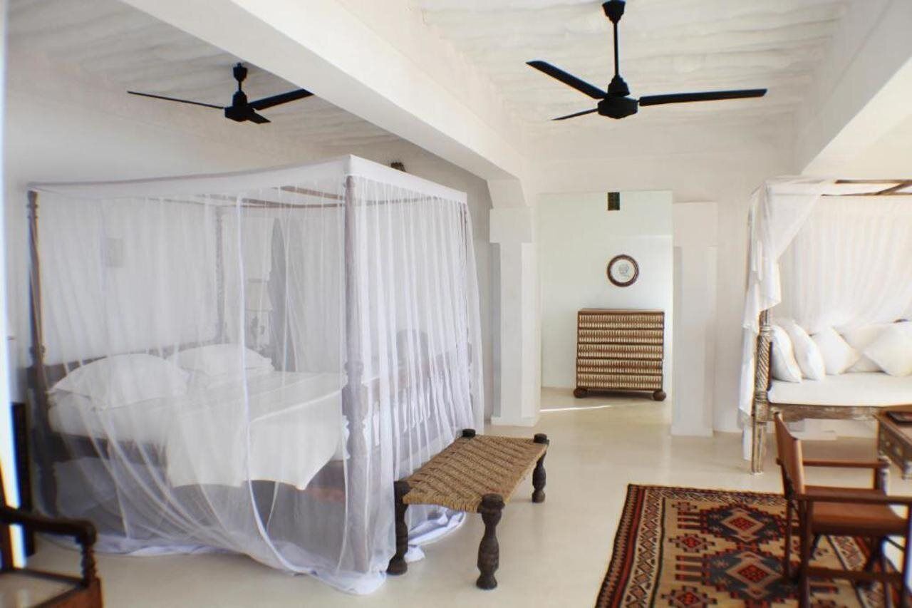 A bedroom with a four-poster bed, a bench, a rattan chair, and a ceiling fan.