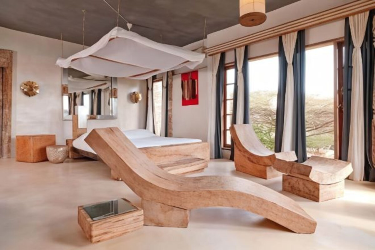 A modern bedroom with a large bed, wooden loungers, and a large window with curtains.