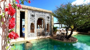 Un espace aménagé au bord de la piscine, avec un bâtiment en pierre, un arbre et une piscine à l’eau d’un bleu limpide.