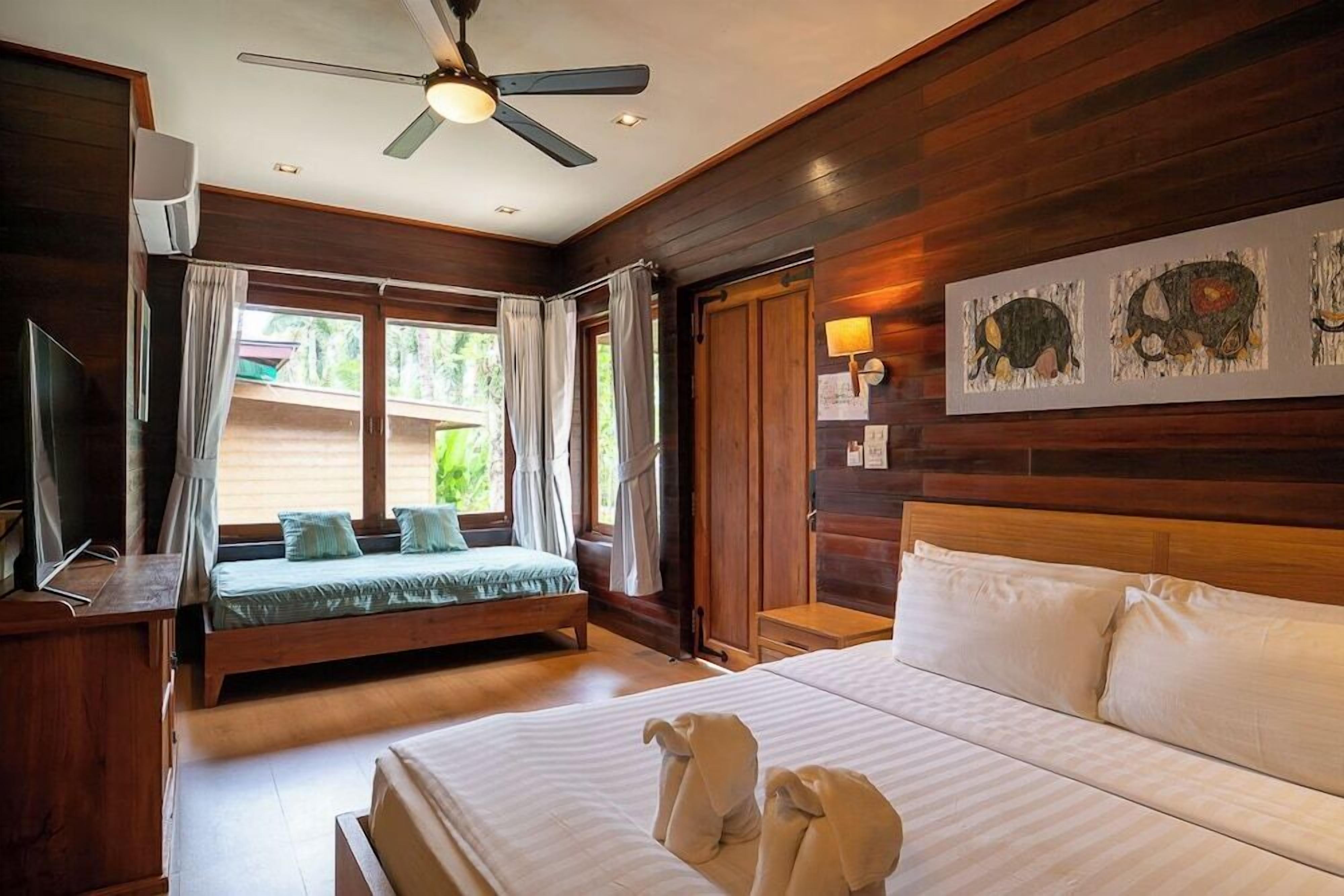 Ein Hotelzimmer mit einem großen Bett, einem kleineren Bett, einem Deckenventilator, einem Fernseher und einem Fenster mit Vorhängen.