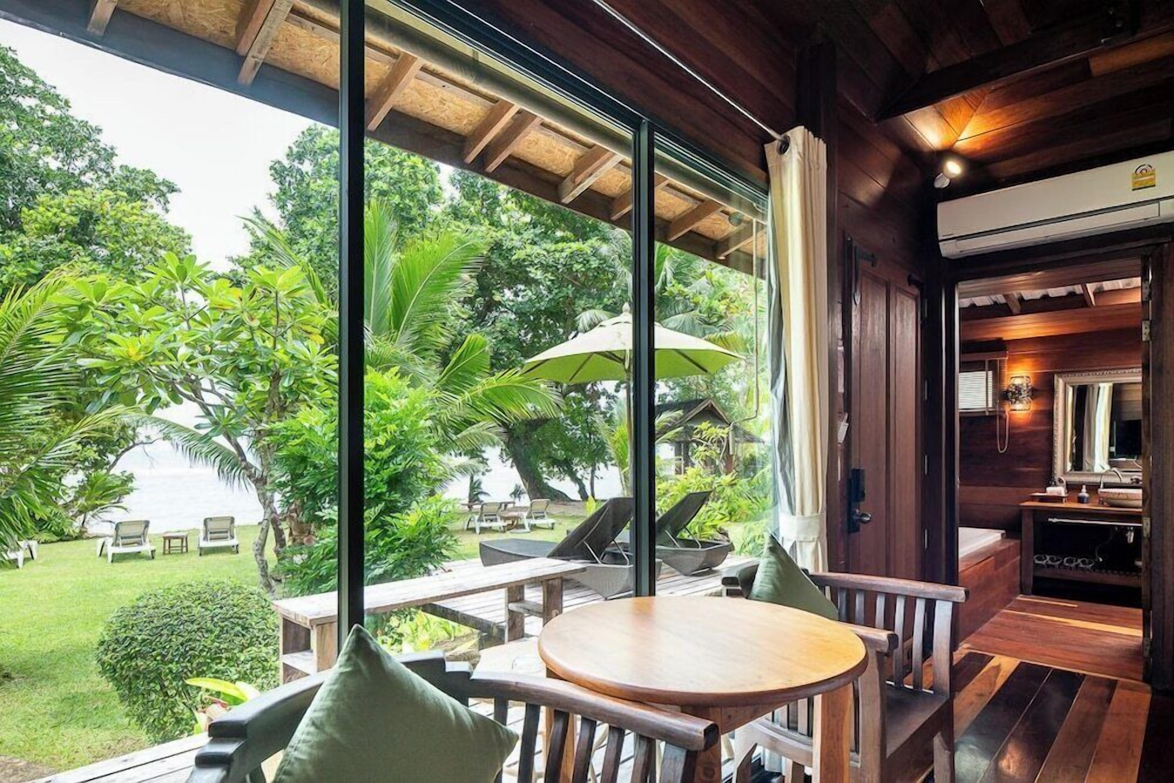 Terraza de madera con una mesa redonda y sillas, con vistas a un jardín exuberante y una zona de piscina.