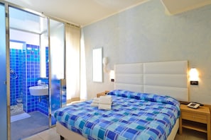 A hotel room with a bed, a bedside table, a nightstand, a wall-mounted phone, and a bathroom with a sink and blue-tiled walls. - Vannucci Hotel (Rimini)