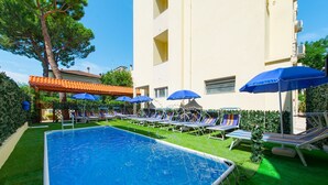 Une piscine entourée de chaises longues et de parasols, elle-même bordée d’une pelouse bien entretenue et d’un bâtiment doté de balcons.
