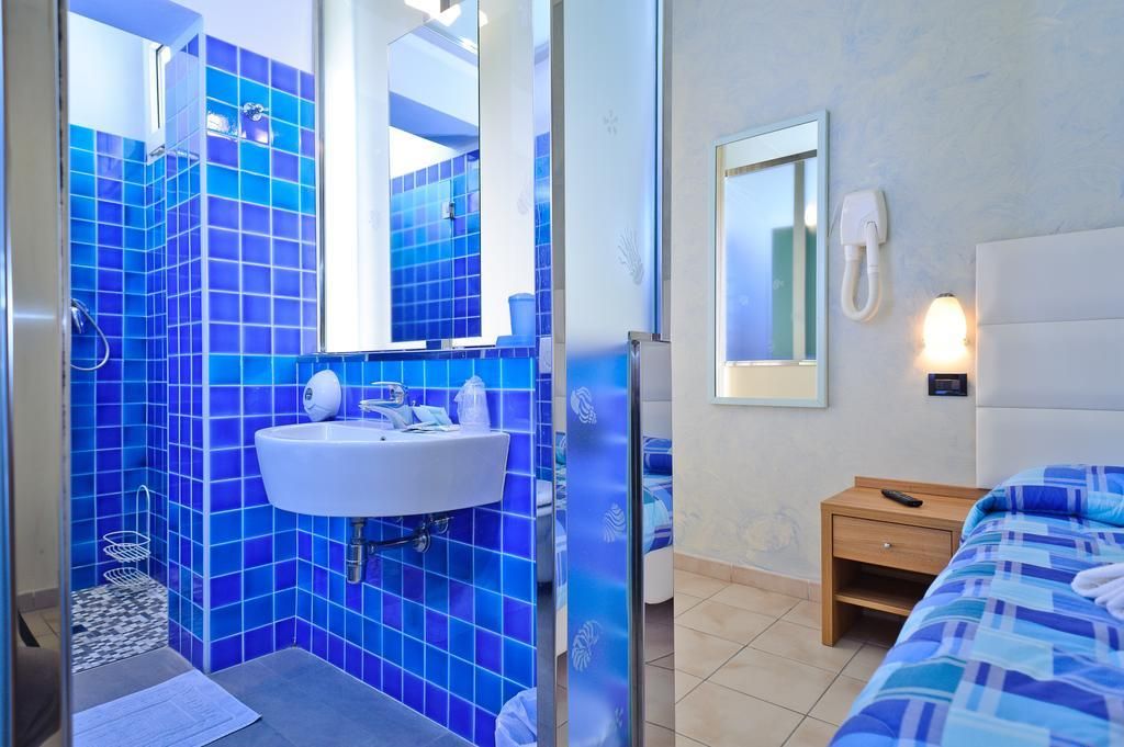 A hotel room with a bathroom featuring blue tiles, a sink, a mirror, and a bedside table with a phone.
