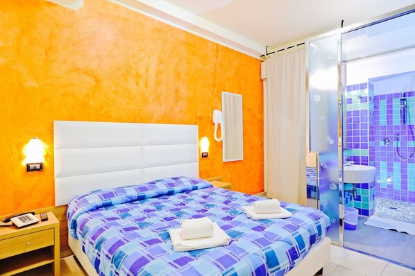 A hotel room with a large bed, a bedside telephone, a nightstand, and a bathroom with a glass shower enclosure. - Vannucci Hotel (Rimini)