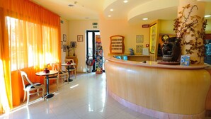 Un hall d’hôtel comprenant un comptoir d’accueil, un petit coin salon avec des tables et des chaises, et une grande fenêtre ornée de rideaux orange.