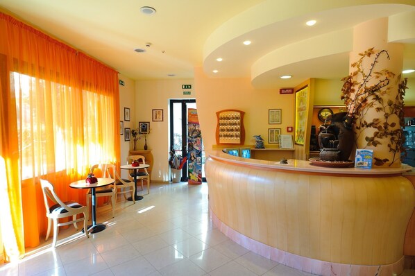 A hotel lobby with a reception desk, a small seating area with tables and chairs, and a large window with orange curtains.