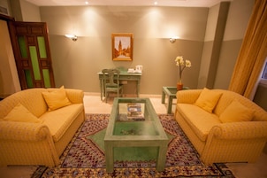 Una sala de estar con dos sofás amarillos, una mesa de centro de vidrio, un tapete estampado, un cuadro en la pared y un escritorio con una silla.