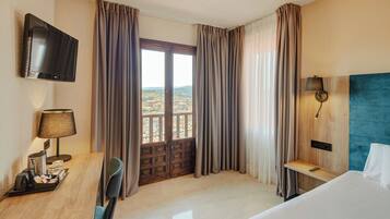 Habitación de hotel con cama, escritorio con lámpara, televisión y vista a la ciudad a través de un ventanal grande.