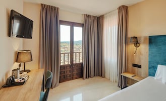 Habitación de hotel con cama, escritorio con lámpara, televisión y vista a la ciudad a través de un ventanal grande.