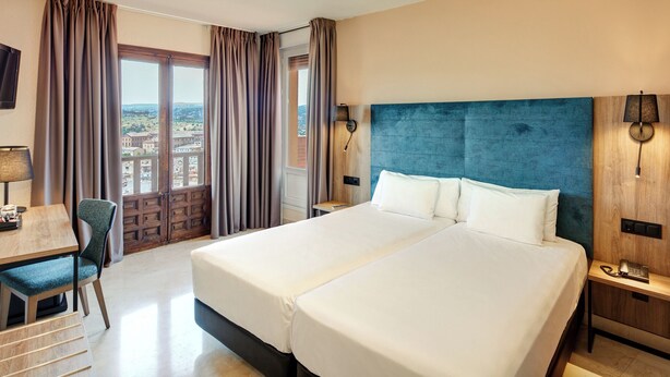 A hotel room with a large bed, a desk with a chair, a TV, and a view of the city through the window. - Sercotel Alfonso Vi (Toledo)