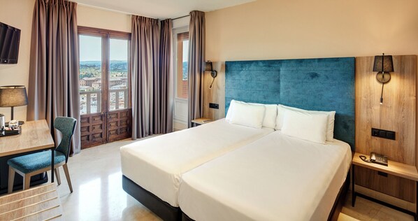 A hotel room with a large bed, a desk with a chair, a TV, and a view of the city through the window. - Sercotel Alfonso Vi (Toledo)