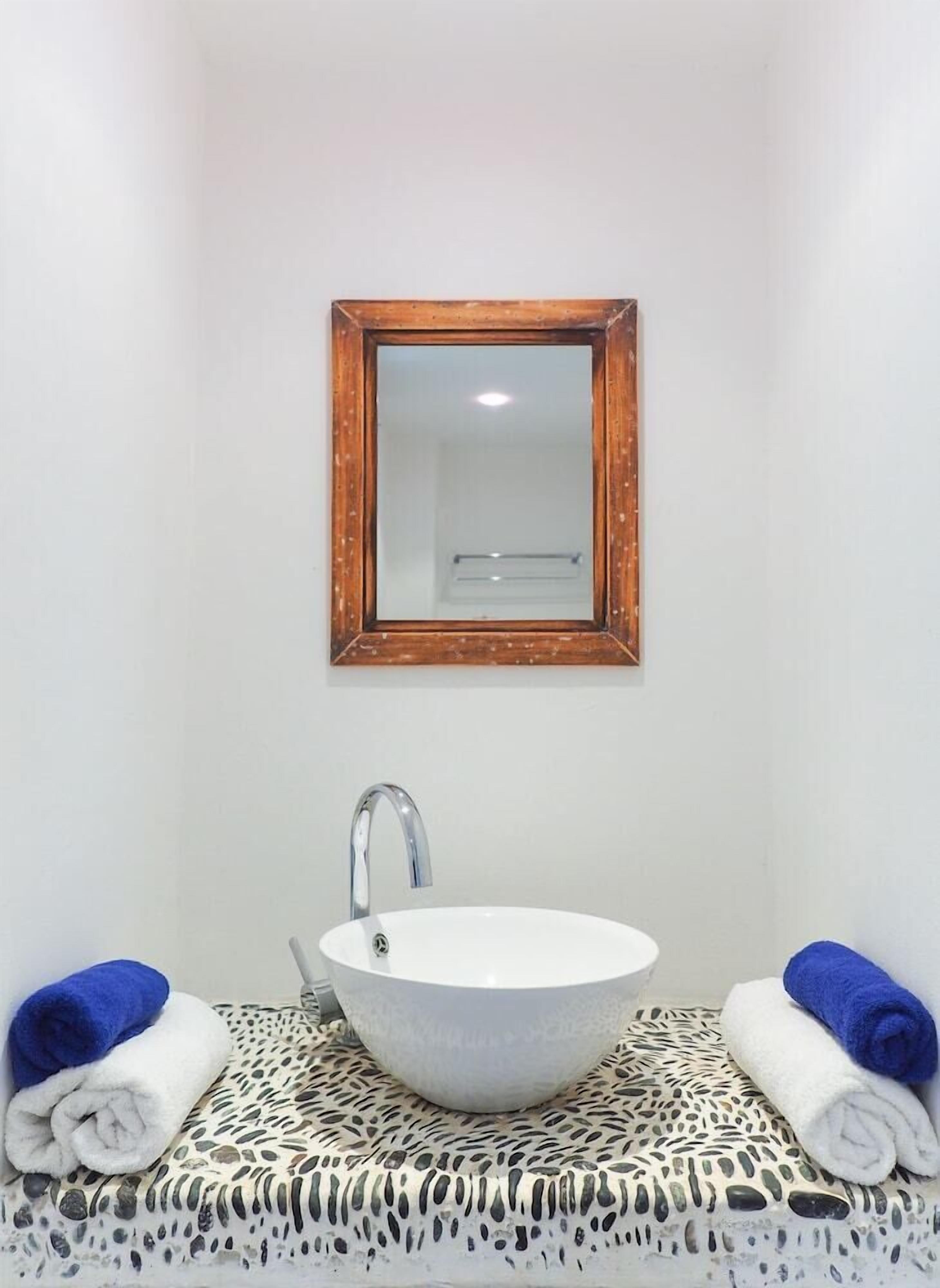 A bathroom with a white sink, a wooden-framed mirror, and two towels on a patterned mat.