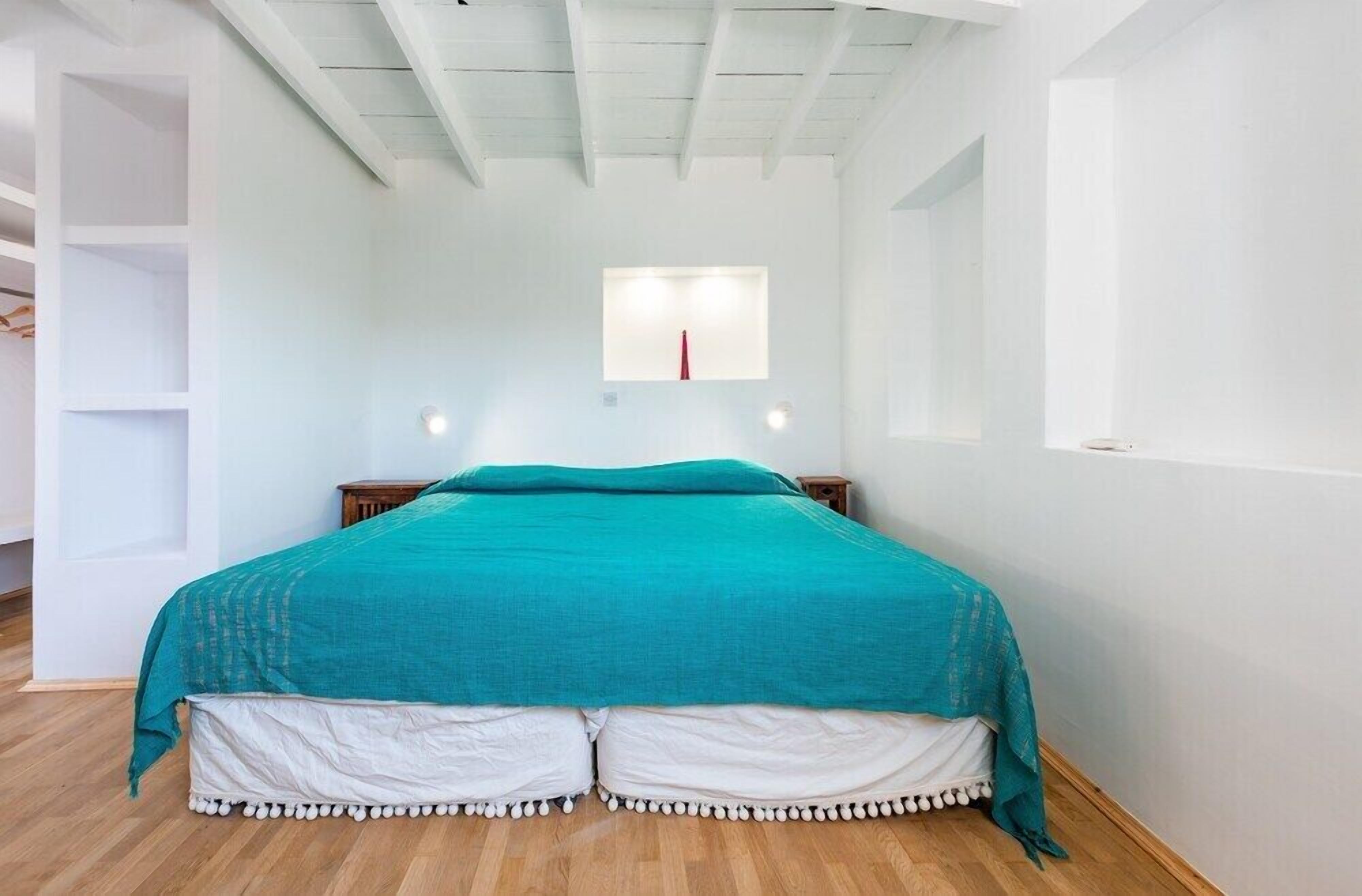 A bedroom with a teal bedspread, white pillows, and a white skirted bed base. There is a small window with a red curtain, a white wall, and a wooden floor.