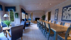 Un vaste espace de salle à manger avec une longue table en bois et des chaises bleues, un coin salon avec des canapés et une lampe, et une grande fenêtre avec des rideaux.