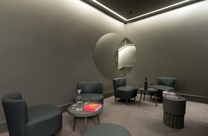 Una habitación moderna con poca luz, un espejo grande, tres sillones verde oscuro, una mesita redonda con un libro y una botella, y un otomanillo.
