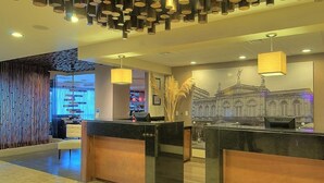 A hotel reception area with a modern design, featuring a reception desk, a decorative wall with a classical building illustration, and a unique ceiling light fixture. - Tryp San Jose Sabana (San José)