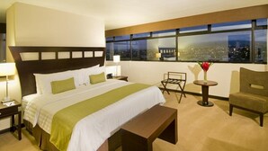 A hotel room with a large bed, a bench, a small table, a chair, and a view of the city at night. - Tryp San Jose Sabana (San José)