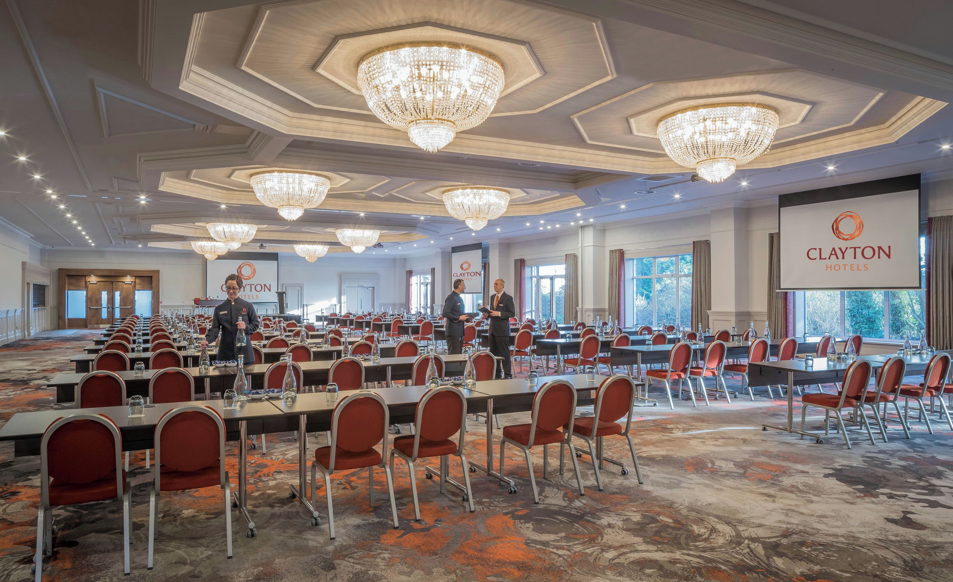 A conference room with tables, chairs, chandeliers, and a large screen displaying the Clayton Hotels logo.