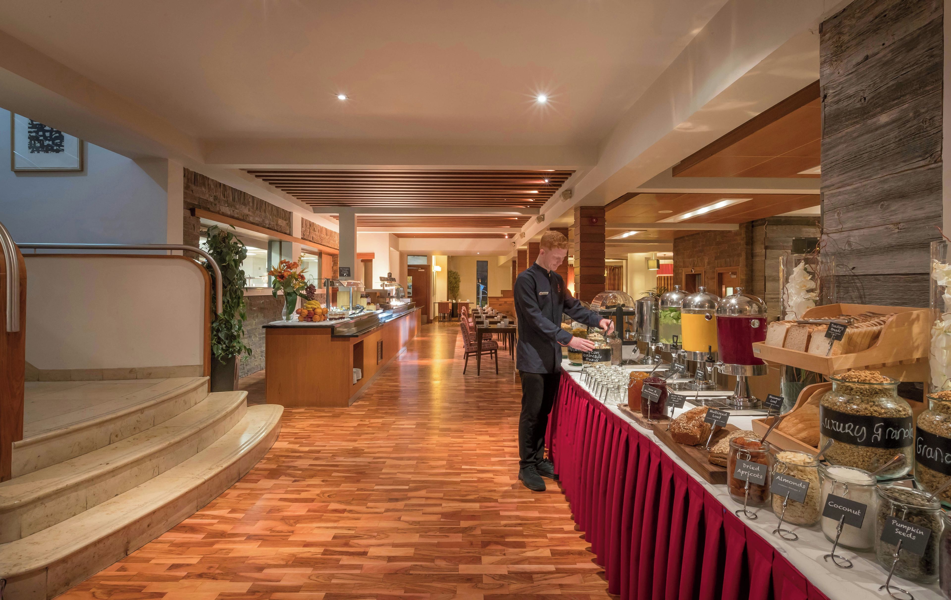 A buffet area with a variety of food options, a barista preparing drinks, and a staircase leading to an upper level.