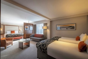 A hotel room with a bed, two sofas, a chair, a glass coffee table, and a painting on the wall.
