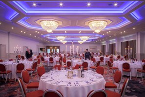 Un salón de banquetes con mesas redondas, sillas rojas, candelabros y suelo alfombrado.