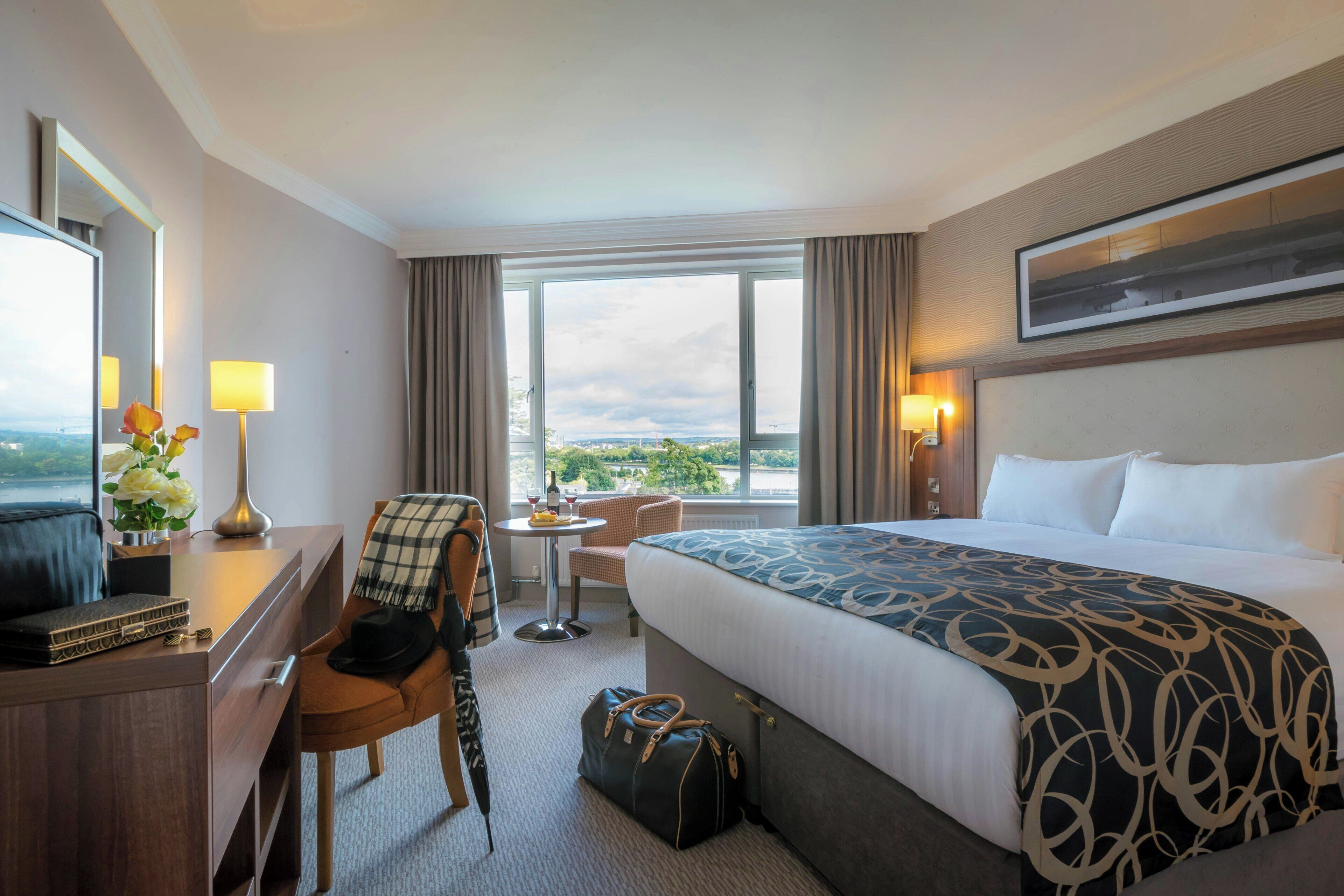 A hotel room with a large bed, a desk with a chair, a small table with a lamp, a suitcase, and a view of the city through the window.