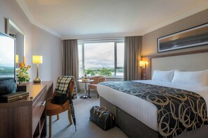 A hotel room with a large bed, a desk with a chair, a small table with a lamp, a suitcase, and a view of the city through the window. - Clayton Hotel Silver Springs (Cork)
