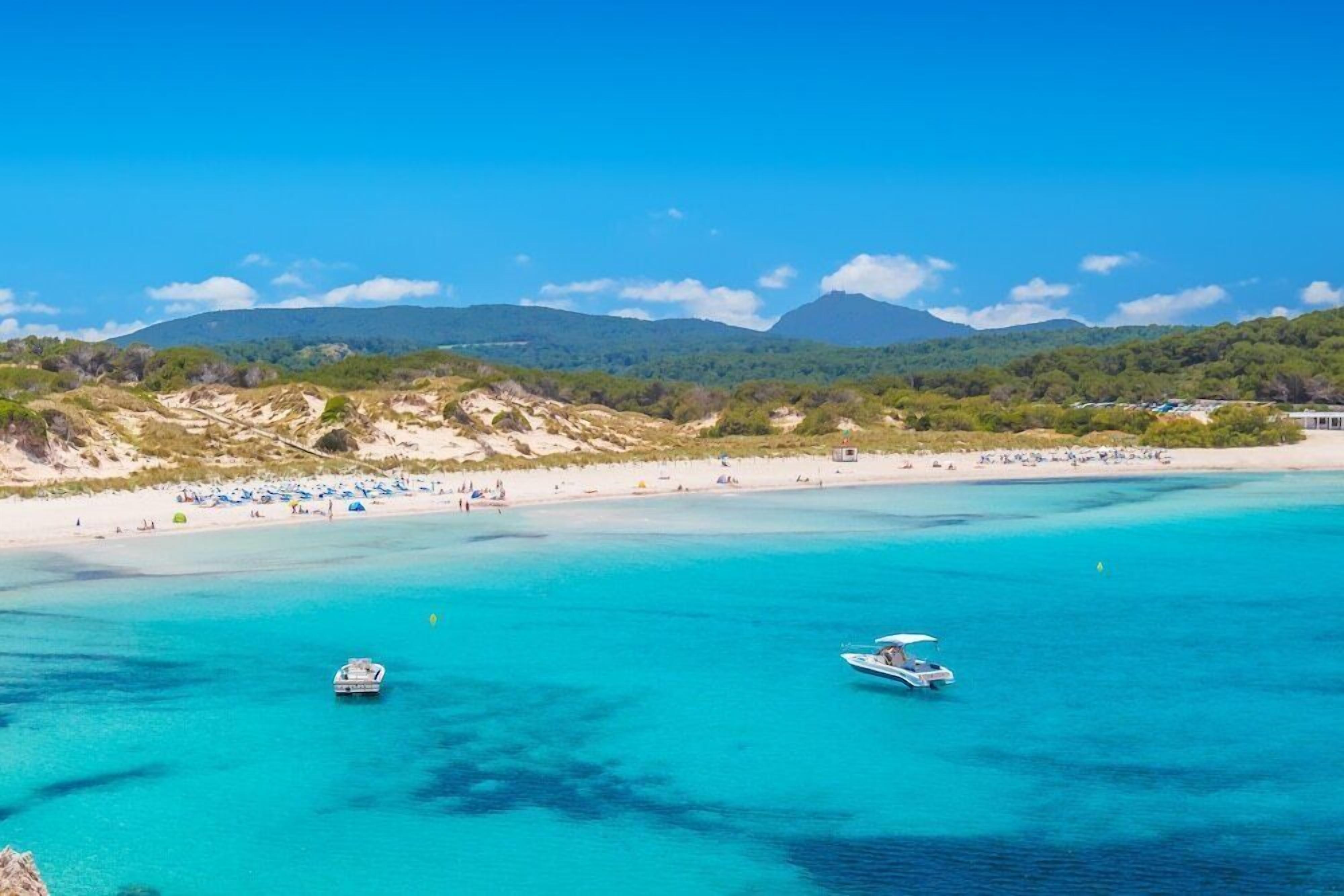 A beach with clear turquoise waters, boats, and a sandy shore with umbrellas.