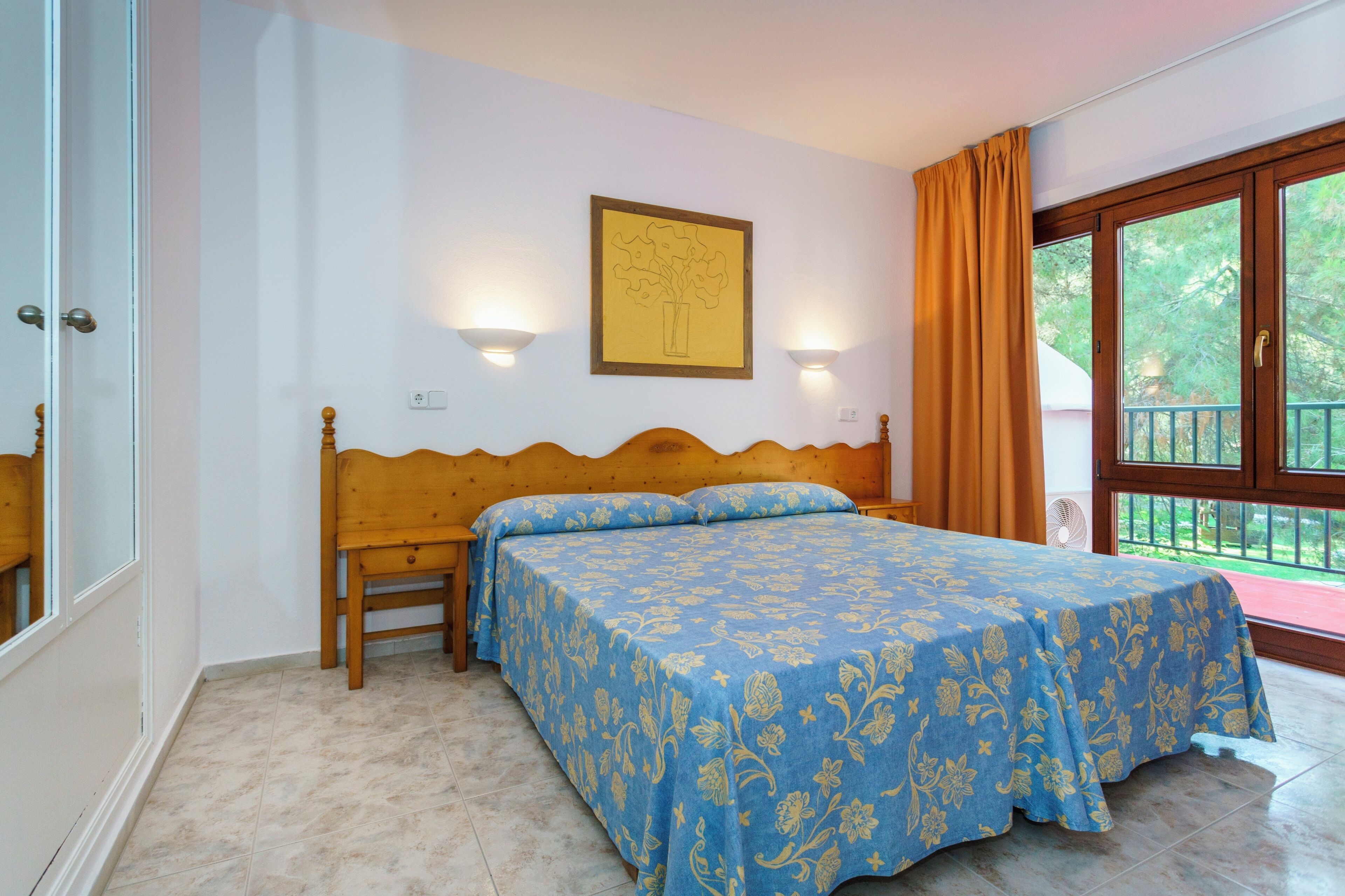 A bedroom with a wooden bed, a blue floral bedspread, two wall-mounted lights, and a window with curtains.