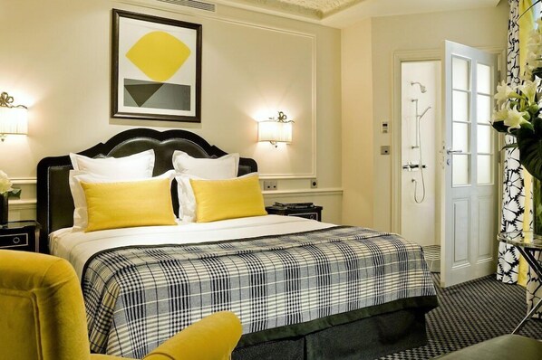 Una habitación de hotel con una cama grande, dos lámparas de noche, un cuadro enmarcado en la pared y una puerta de baño visible al fondo.