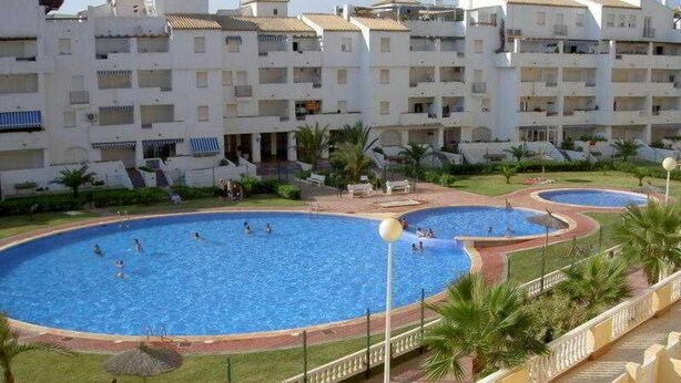 Un espace piscine comprenant plusieurs bassins, entouré d’un bâtiment doté de balcons et d’une pelouse bien entretenue.