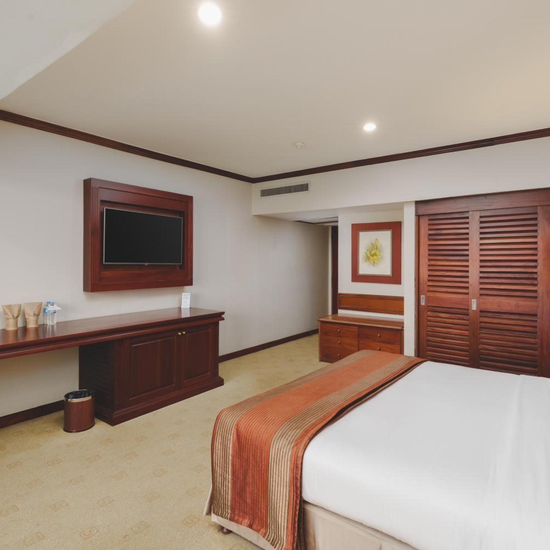 A hotel room with a bed, a TV mounted on the wall, a desk with a chair, a wardrobe, and a small trash bin.