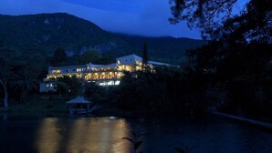 A large, multi-story building with a prominent entrance and a covered area near a body of water, surrounded by dense vegetation and hills. - Hunas Falls Hotel Kandy (Kandy)