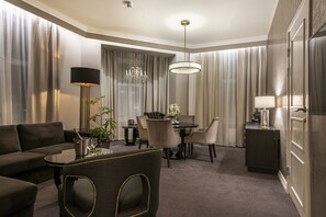 Ett hotellrum med en soffa, ett matbord, stolar, en kristallkrona och ett skrivbord med en lampa.