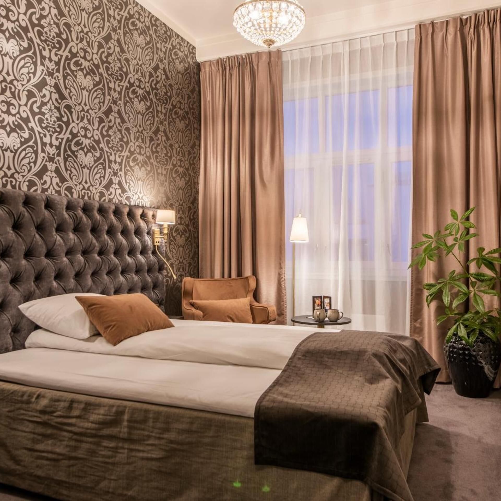 A bedroom with a tufted headboard, a bed with brown bedding, a side table with a lamp, and a potted plant.