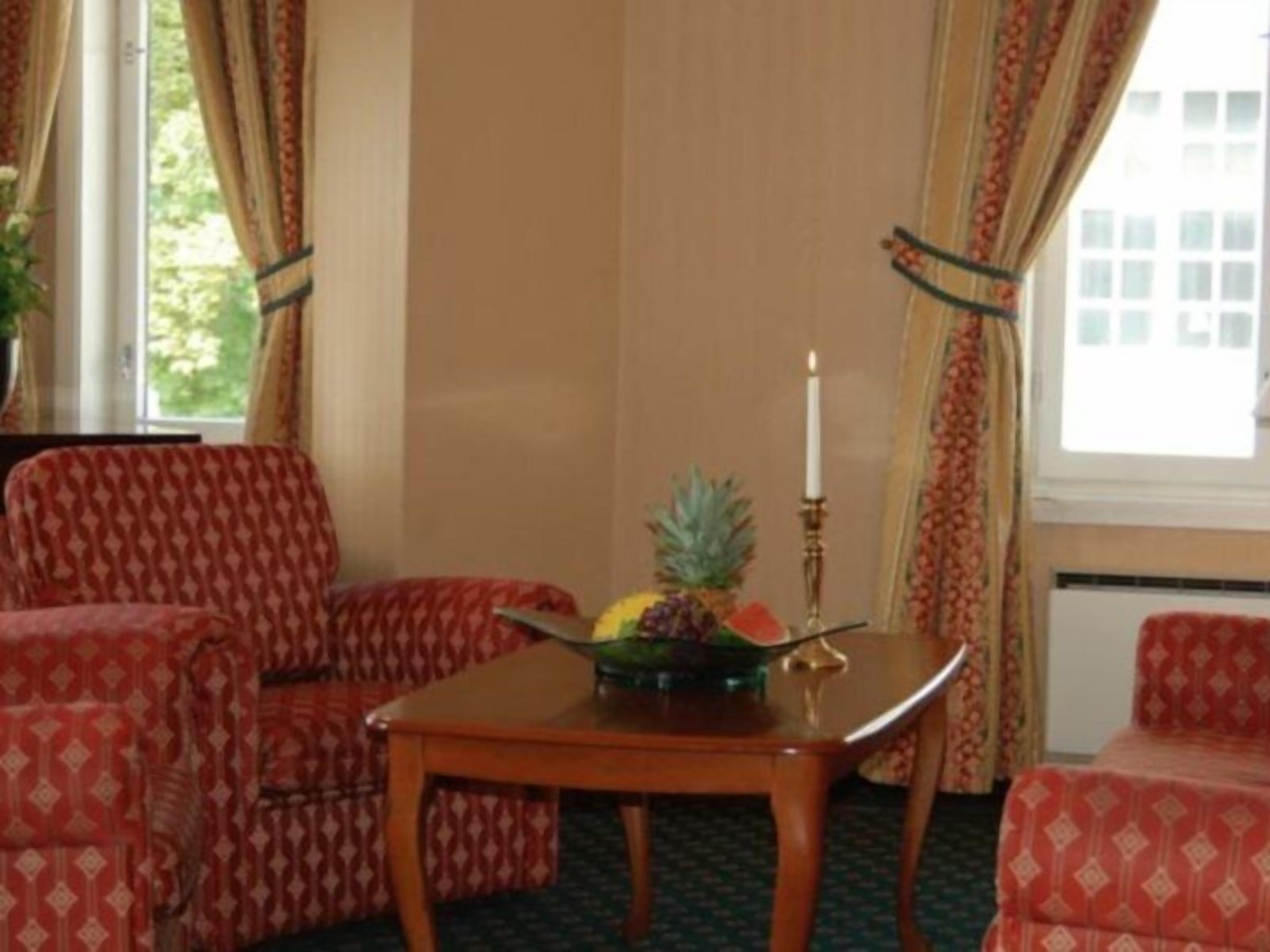 Una habitación con un sofá rojo estampado, una mesa de madera con un frutero y una vela, y una ventana con cortinas.
