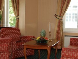 Una habitación con un sofá rojo estampado, una mesa de madera con un frutero y una vela, y una ventana con cortinas.