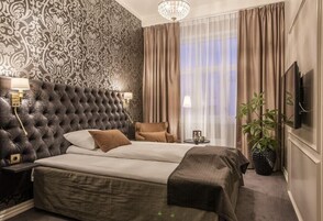 Un dormitorio con cabecera capitonada, una cama con dos almohadas, una mesita de noche con lámpara, una silla, una planta en maceta y un televisor en la pared.