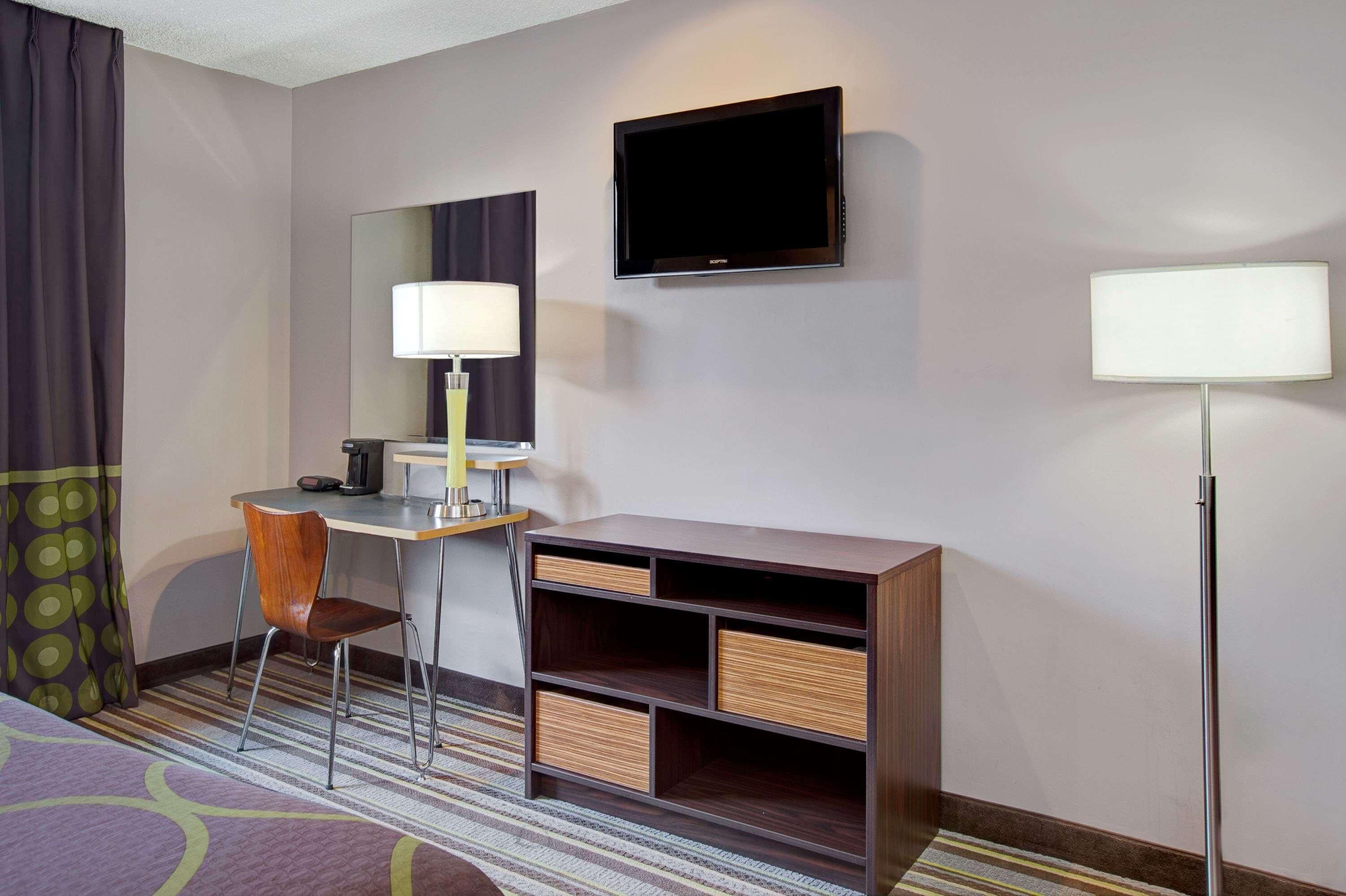 A hotel room with a bed, a desk with a chair, a TV, a lamp, and a bookshelf.