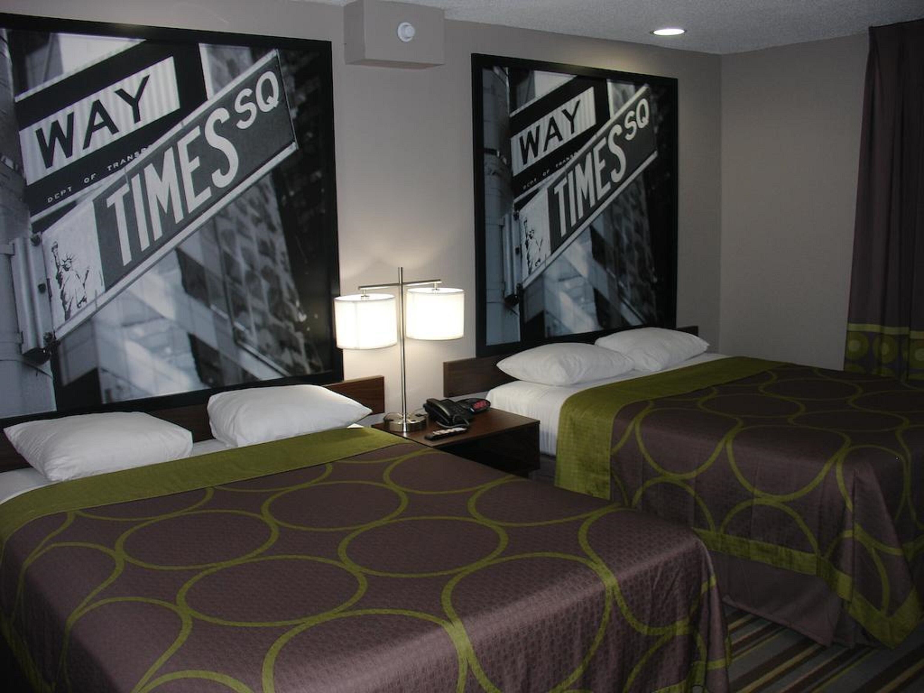 A hotel room with two beds, a nightstand, a lamp, and two large posters on the wall.
