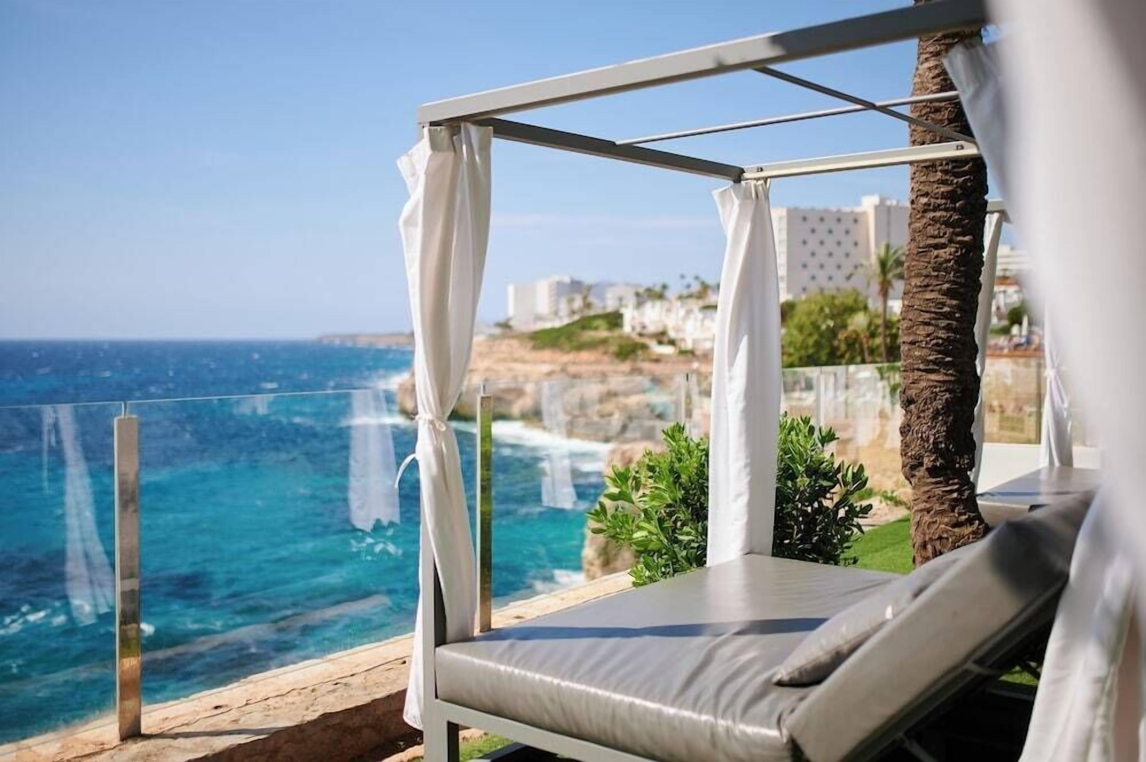Ein Balkon am Strand mit einer gepolsterten Liege, einer Glastrennwand und Blick auf das Meer und entfernte Gebäude.