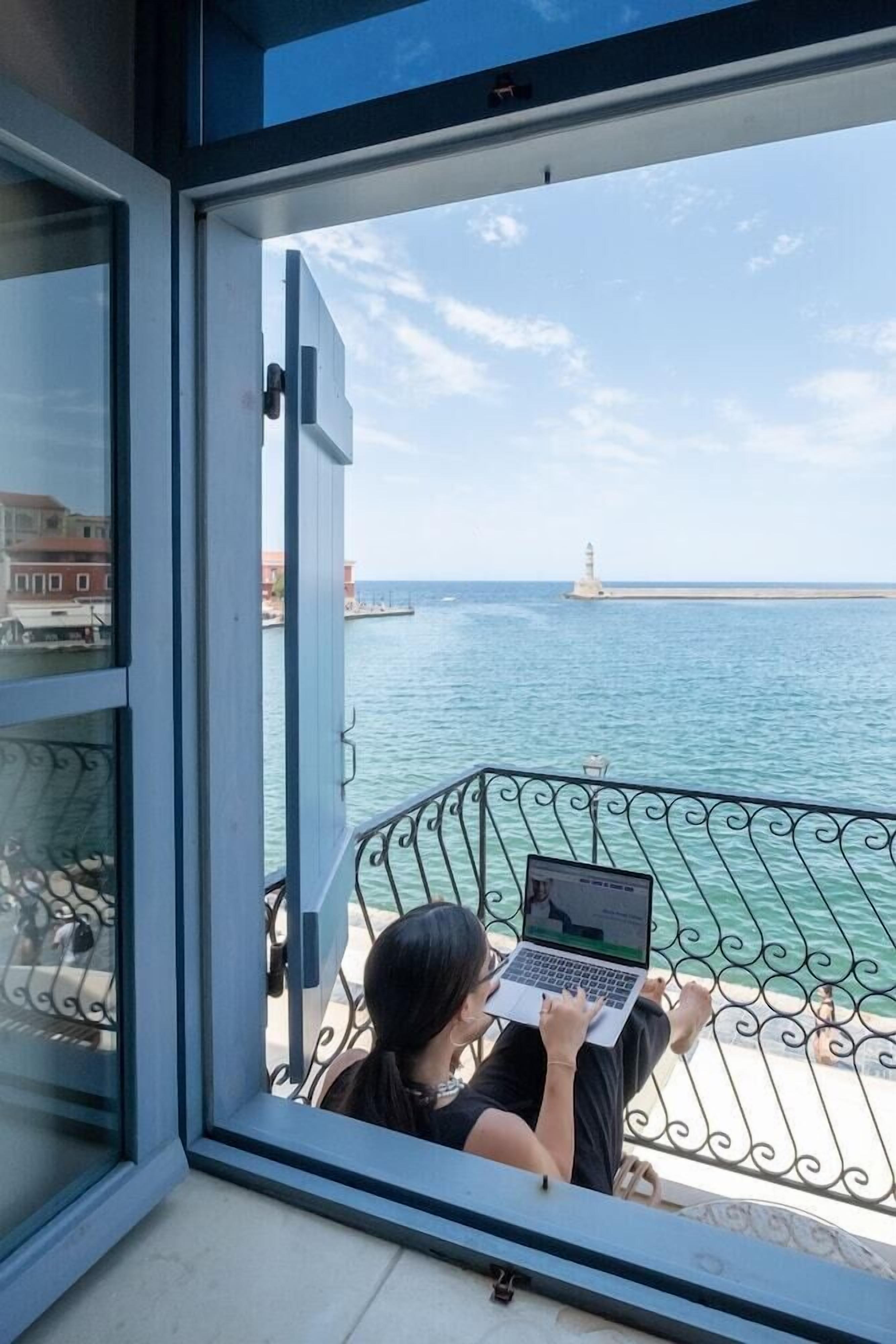 A person using a laptop on a balcony with a view of the sea and a lighthouse.