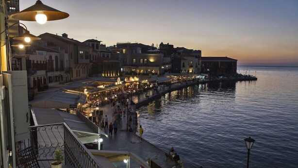 A waterfront promenade with buildings, a pier extending into the water, and people walking along the path.