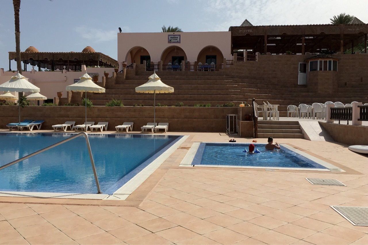 Ein Poolbereich mit zwei Schwimmbecken, Liegestühlen, Sonnenschirmen und einem Gebäude mit Bögen im Hintergrund.