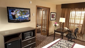Ein Hotelzimmer mit Flachbildfernseher, der eine Wettervorhersage zeigt, einem Schreibtisch mit Lampe, einem Sessel und einem Bett mit gemustertem Überwurf.