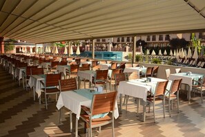 A covered outdoor dining area with tables and chairs, a pool in the background, and a view of a building with balconies. - NOX INN Club Hotel (Alanya)