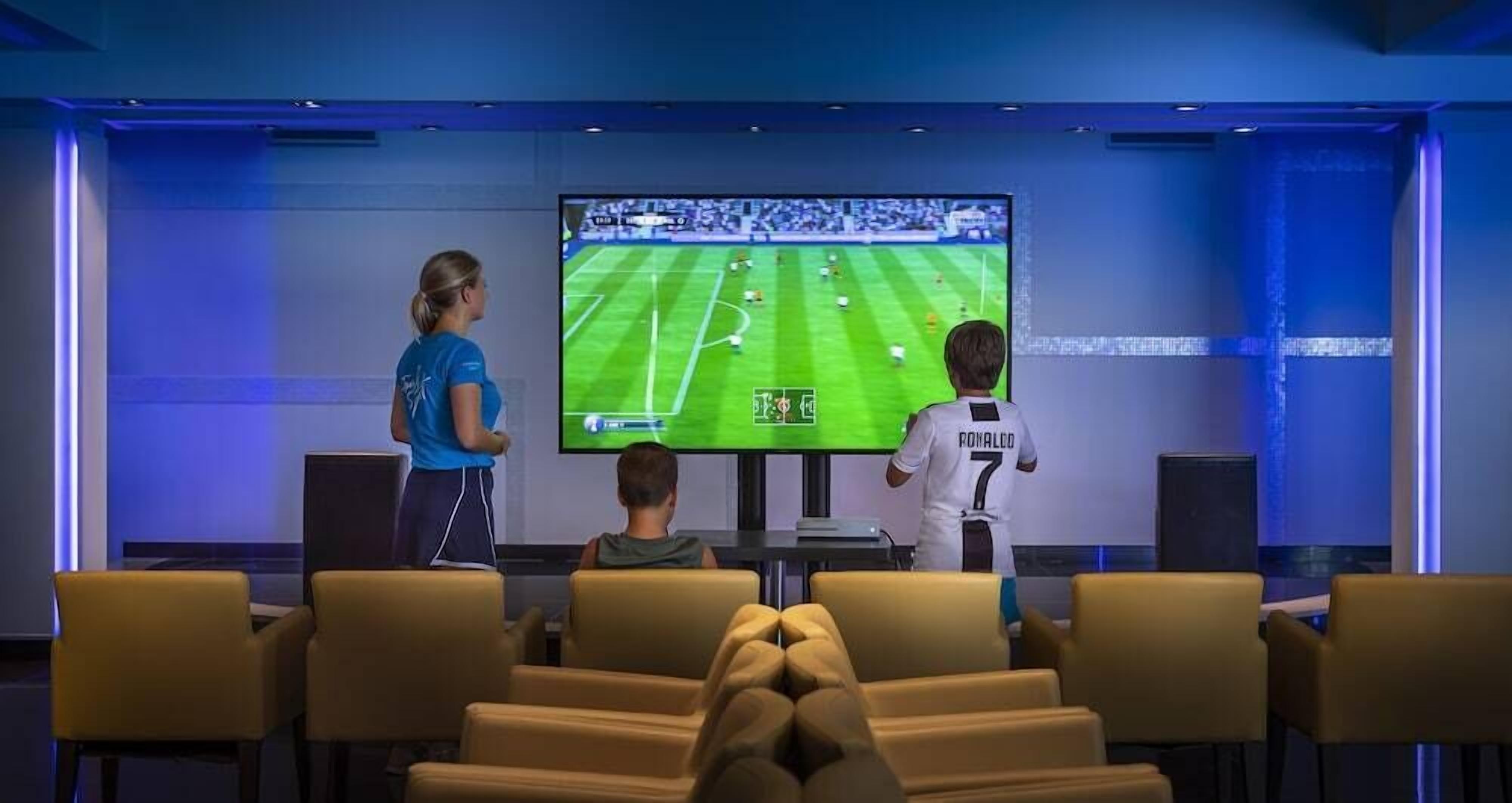 Una habitación con una pantalla grande mostrando un partido de fútbol, un niño y un adulto observando, y un área de descanso con sillones.