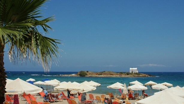 A beach with white umbrellas and orange chairs, a palm tree, and a small white building on a rocky outcrop. - Comfort Vaggelis Studios (Hersonissos)
